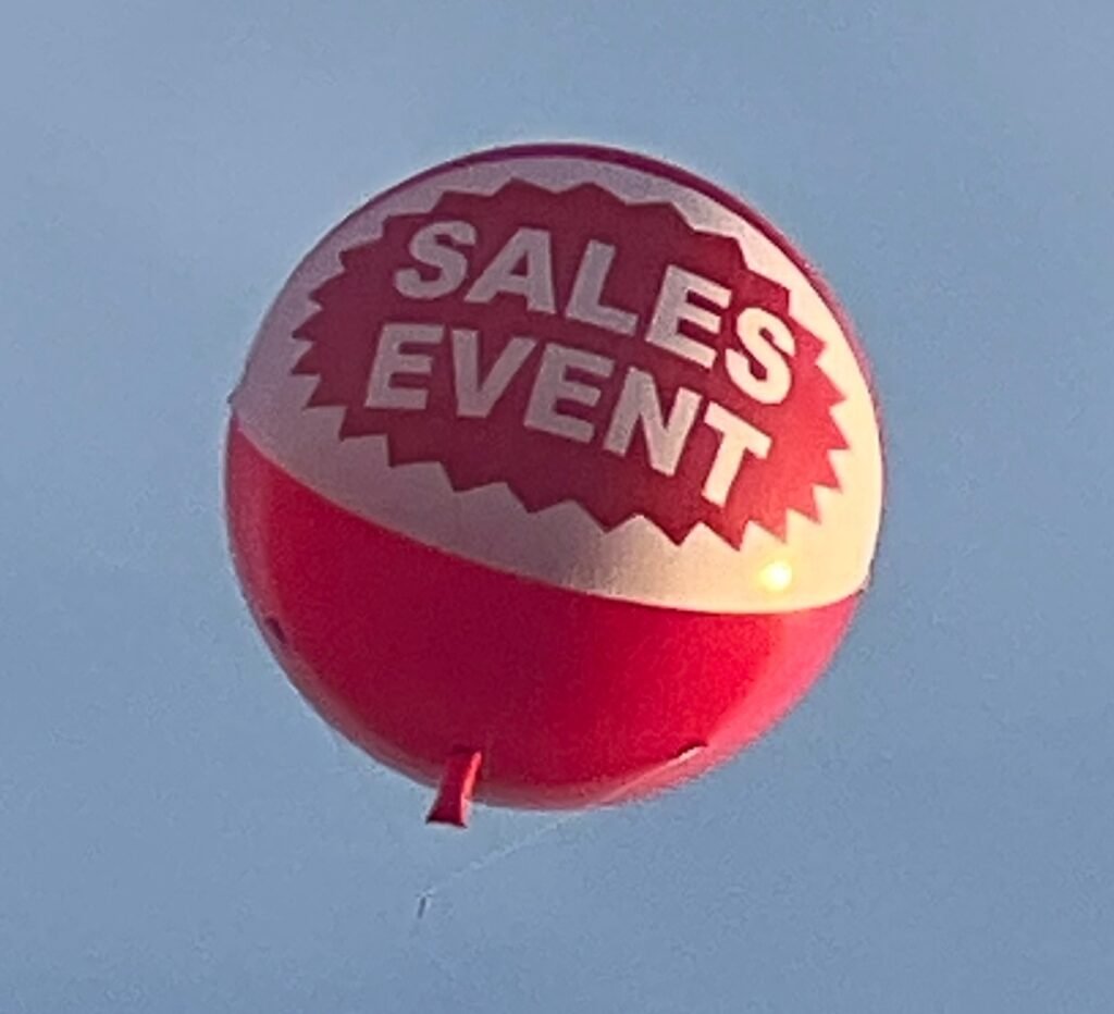 Round red and white SALES EVENT helium advertising balloon flying against a clear sky